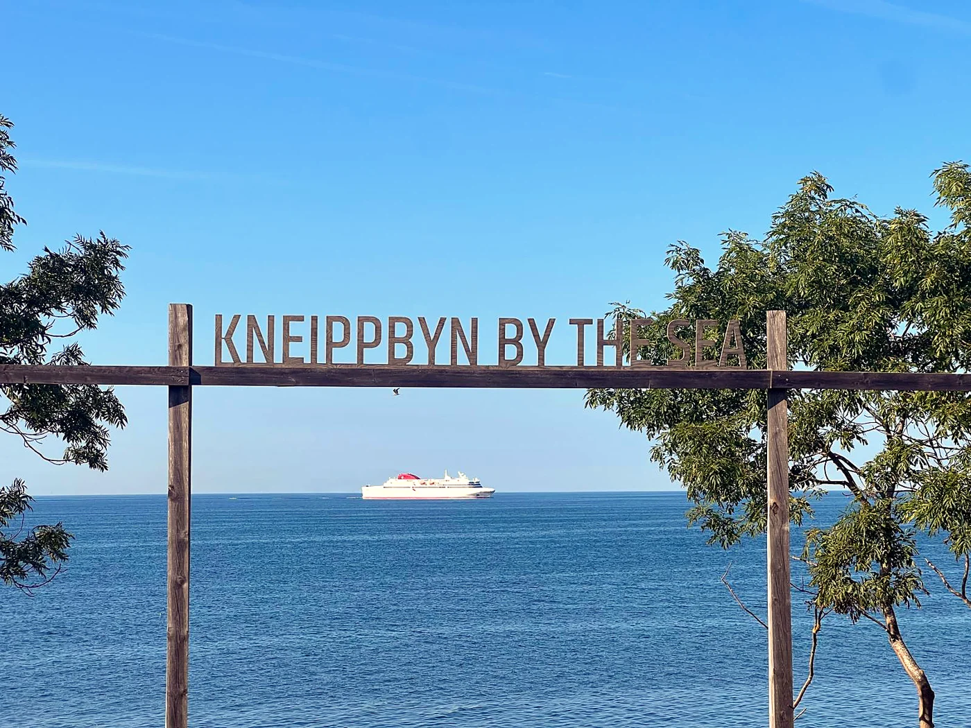 The Kneippbyn sign framing the blue sea and a passing ferry on Gotland, Sweden.